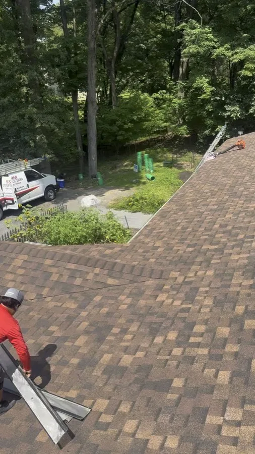 SSKM Construction crew member carrying step flashing along a roof ridge, with the company van parked below