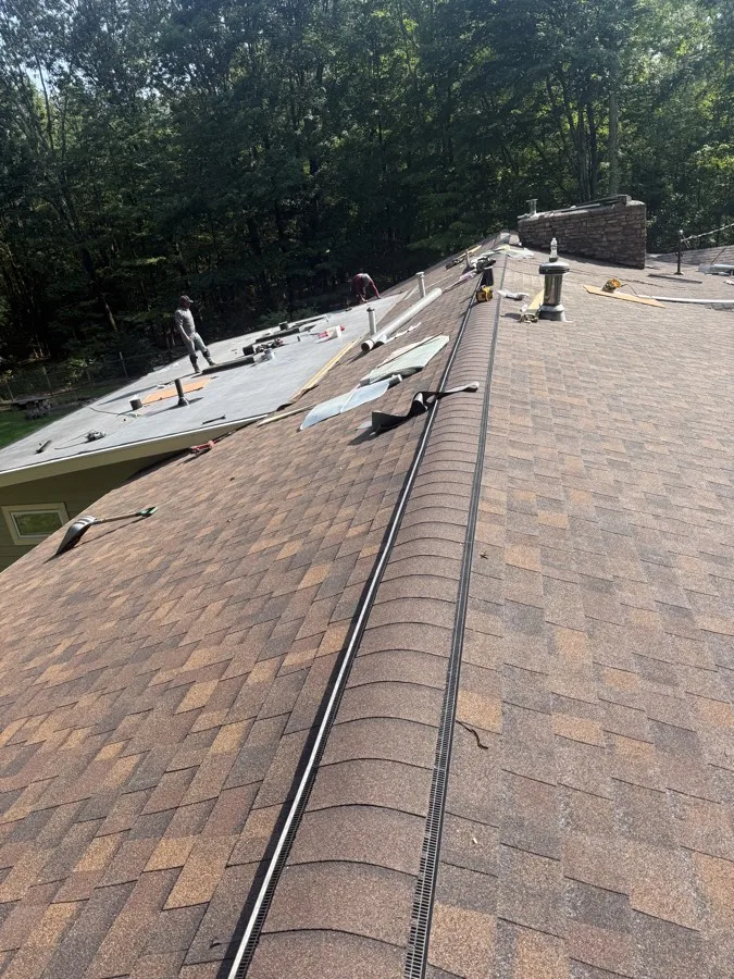 New ridge vent and flashing installed along a shingle roofline, with a flat roof section and stone chimney in the background